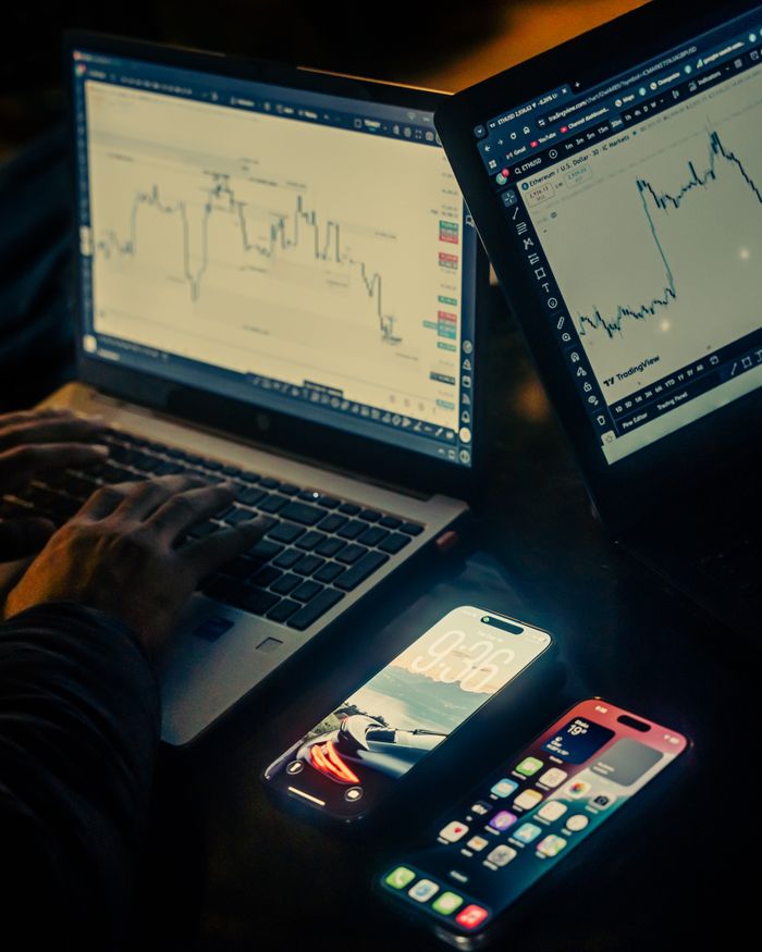 A trading workspace with laptops displaying charts and smartphones, set in a low light environment, Dhaka.