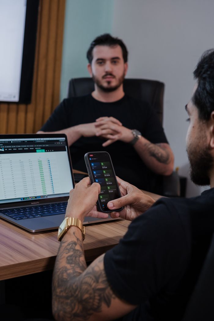Two men in a modern office reviewing stock market data on laptop and smartphone.